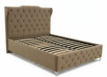 A beige upholstered bed frame with a tall tufted headboard and slatted base, photographed in a furniture showroom with other beds and mattresses visible in the background.