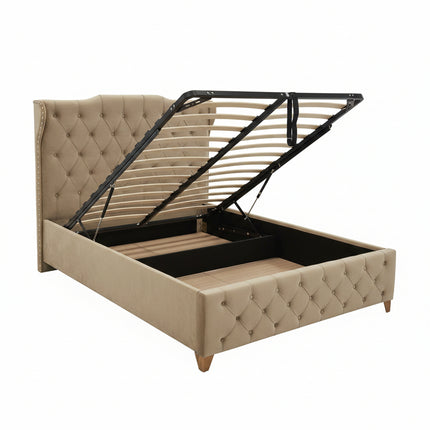 A tufted beige upholstered bed frame with a lift-up headboard and open storage base, photographed in a furniture showroom with other beds and showroom fixtures visible.