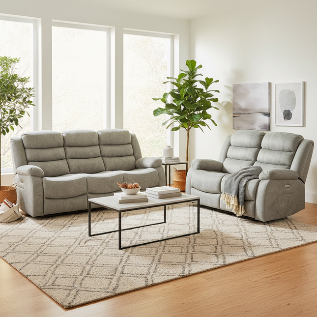 A light grey upholstered living room furniture set in a bright, modern living room. The set includes a three-seat sofa, a matching recliner with a throw blanket, a small black side table, and a large patterned area rug on a light wood floor.