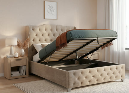 A beige tufted ottoman storage divan bed with the lift-up mattress platform open, styled in a warm beige bedroom with a matching nightstand, lamp, rug, and curtains.