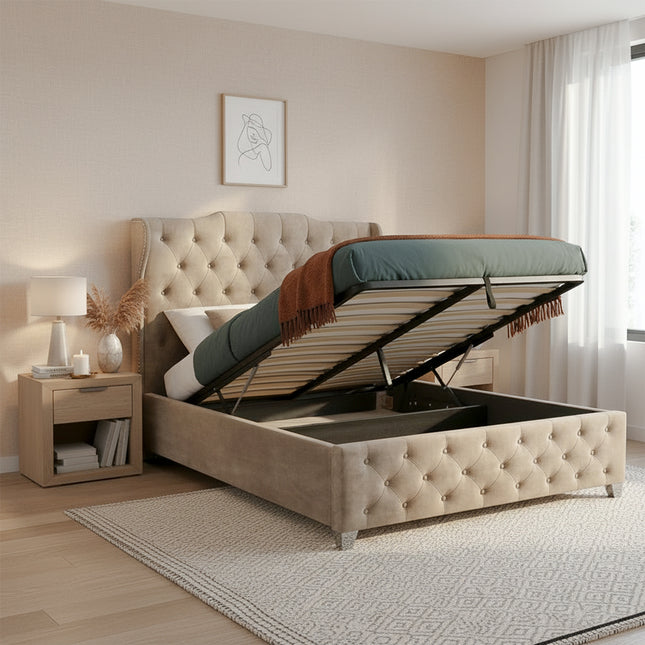 A beige tufted ottoman storage divan bed with the lift-up mattress platform open, styled in a warm beige bedroom with a matching nightstand, lamp, rug, and curtains.