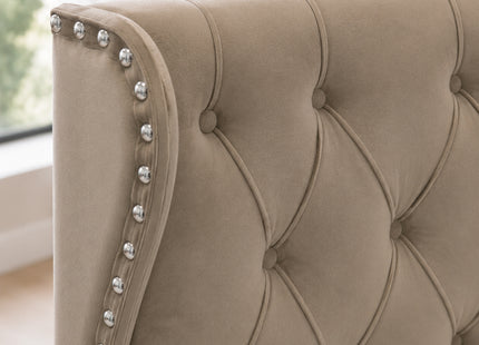 Close-up of a beige tufted upholstered headboard with silver nailhead trim along the curved left edge, shown in a softly lit room.