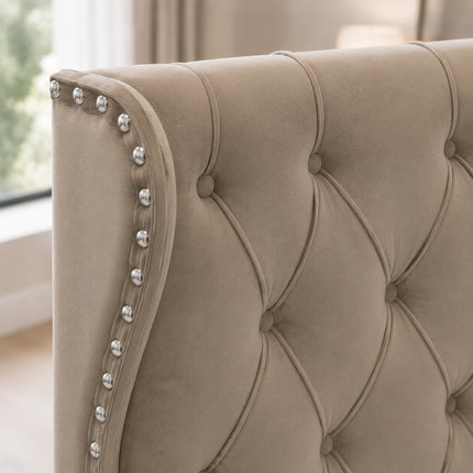 Close-up of a beige tufted upholstered headboard with silver nailhead trim along the curved left edge, shown in a softly lit room.
