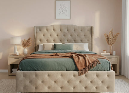 A tufted upholstered ottoman divan bed frame in a warm beige/taupe velvet, styled with sage green bedding and a brown throw blanket in a modern, softly lit bedroom.